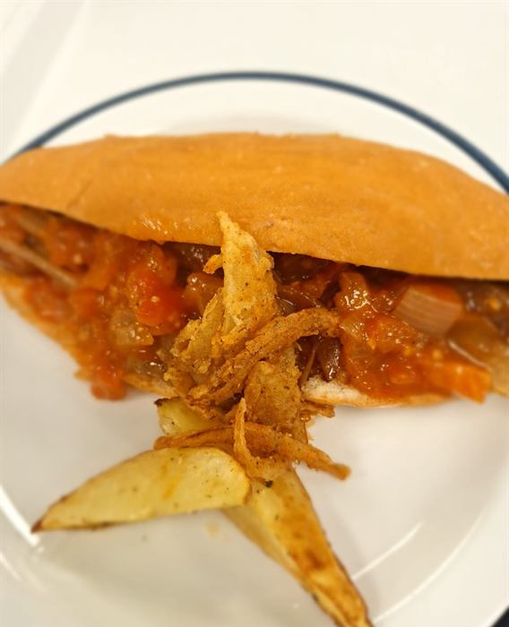 A baguette filled with a tomato and pepper relish, topped with crisp fried onion, with potato wedges